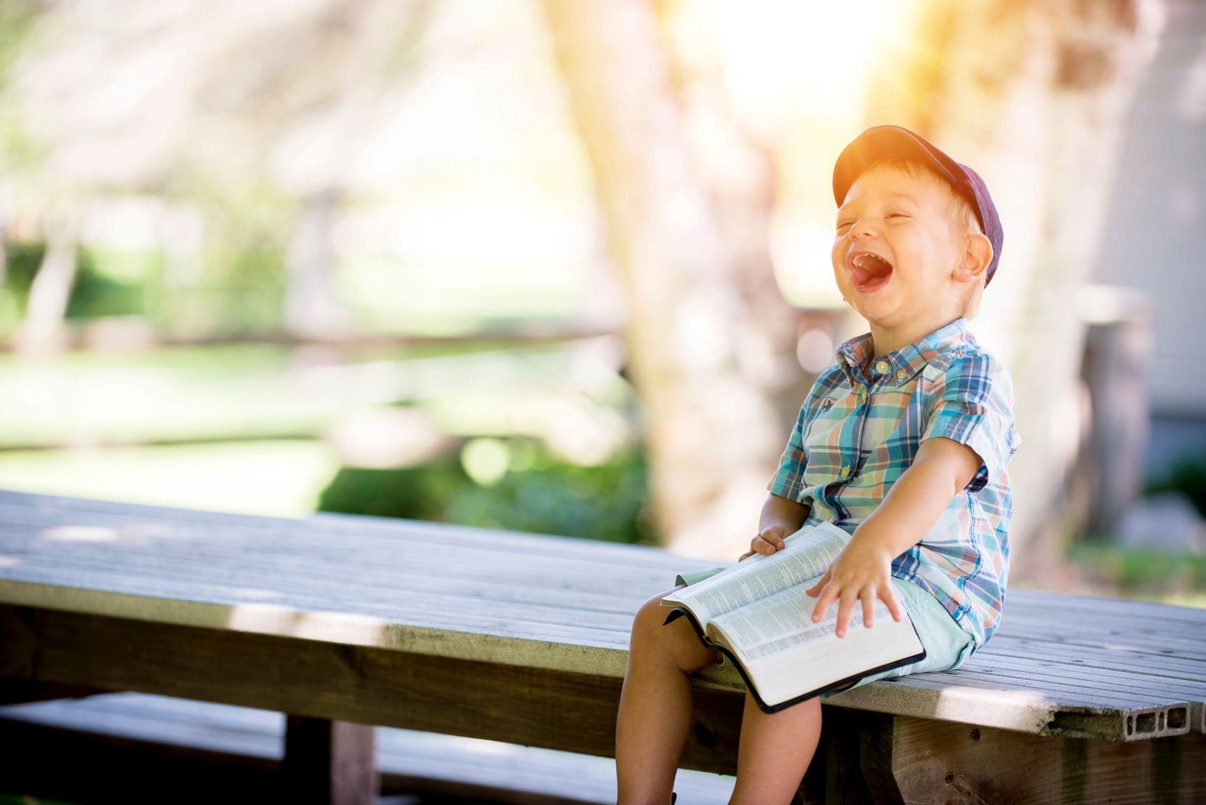 穿着格子衬衫和帽子的孩子坐在外面的长凳上拿着书笑